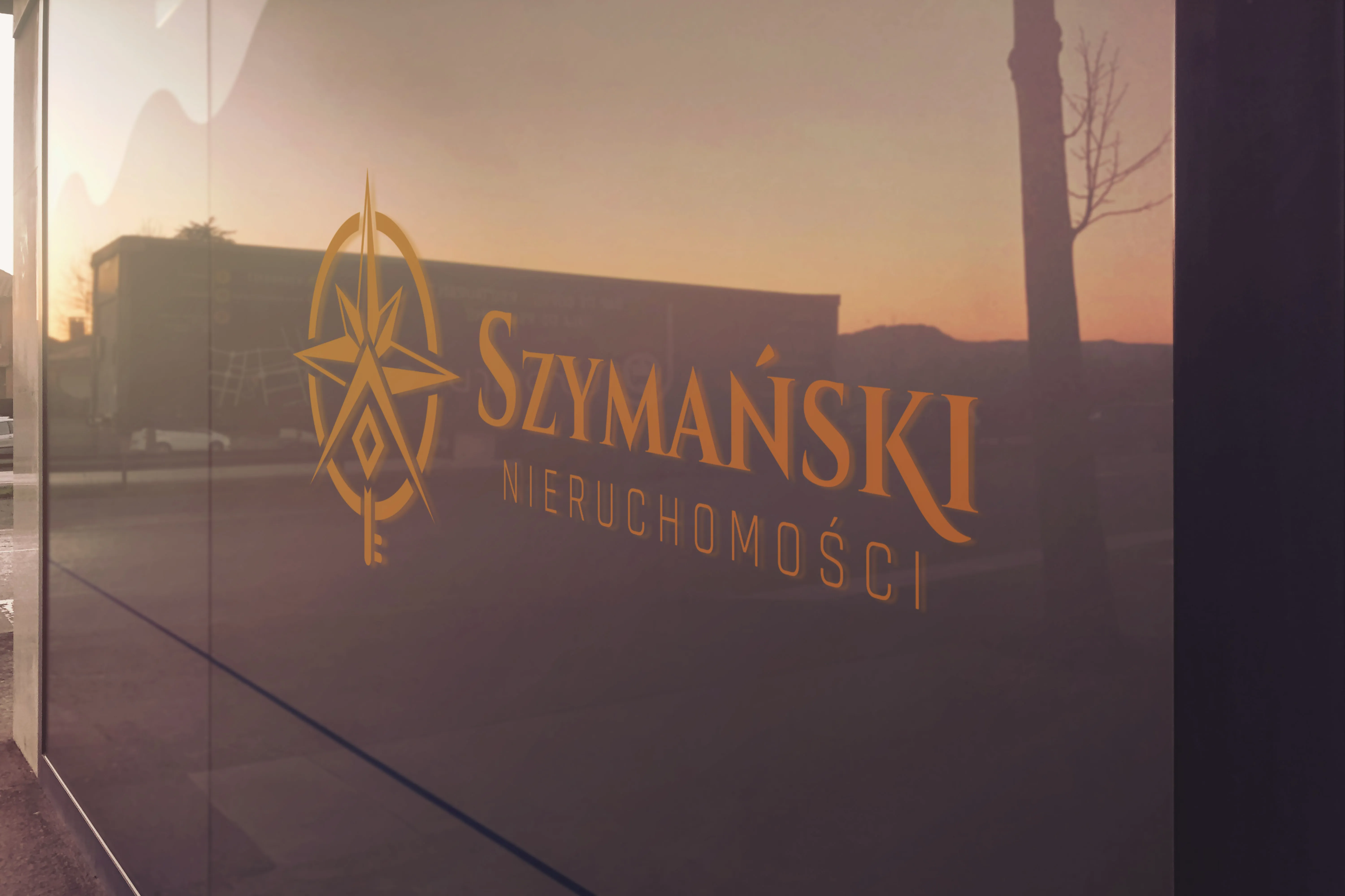 Wizualizacja logo Szymański Nieruchomości na szklanej witrynie biura o zachodzie słońca