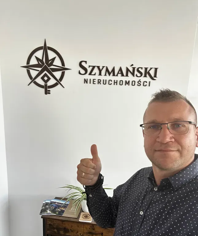 Marek Szymański z uniesionym kciukiem przy znaku Szymański Nieruchomości na ścianie swojego biura
